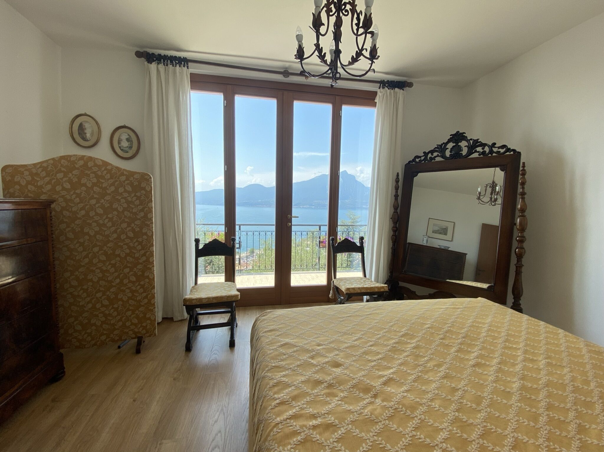 Une chambre avec un grand lit, une commode en bois, une chaise, un miroir et une vue sur la mer, visible à travers une porte coulissante.