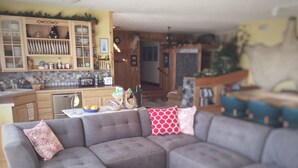 A living room with a sectional sofa, a kitchen area with wooden cabinets, and a dining area with blue chairs. - Pioneer Ridge B&B Inn (Wasilla)