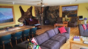 A cozy living room with a stone fireplace, a sofa with red pillows, a dining area with a wooden table and chairs, and a large painting on the wall. - Pioneer Ridge B&B Inn (Wasilla)