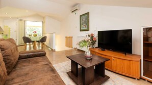 Una sala de estar con un sofá de cuero marrón, una mesa de centro de madera con un jarrón, un televisor de pantalla plana sobre un mueble de madera y una zona de comedor con una mesa y sillas.