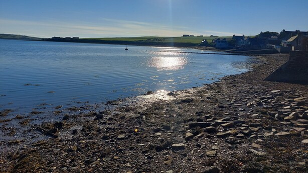 A rocky shoreline with calm water reflecting sunlight, a distant view of houses, and a clear sky.