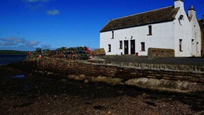 A white building with a stone wall in front, a clear blue sky, and a calm body of water in the background.
