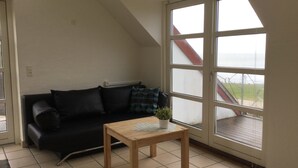 A room with a black leather sofa, a wooden coffee table, and a large window with a view of a red-roofed building. - Holiday home with beautiful sea views. (Frørup)