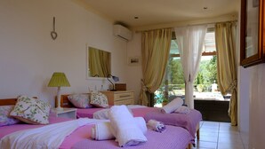 A hotel room with two beds, a window with curtains, a bedside table with a lamp, and a wall-mounted air conditioner.