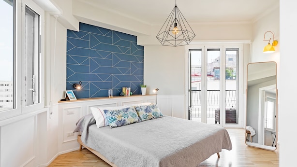 Ein Schlafzimmer mit Bett, einer Wand mit geometrischem Muster, einer Hängelampe und Blick ins Freie.
