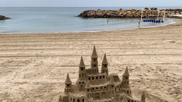 Un grand château de sable doté de plusieurs tours et d’un fossé, situé sur une plage dont le rivage est rocheux.