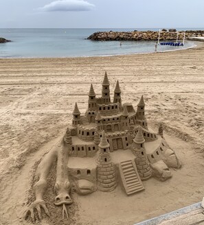 Un grand château de sable doté de plusieurs tours et d’un fossé, situé sur une plage dont le rivage est rocheux.