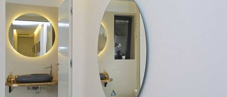 A bathroom with a round mirror, a sink, and a small window with blinds.