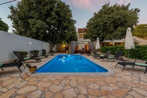 An outdoor swimming pool with lounge chairs and umbrellas, surrounded by stone flooring and trees.