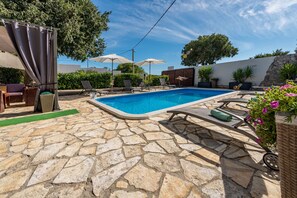 A swimming pool with stone flooring, lounge chairs, umbrellas, and potted plants.