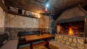 Un intérieur rustique avec un mur en pierre, une table en bois, un banc et une cheminée où l’on peut voir le feu.