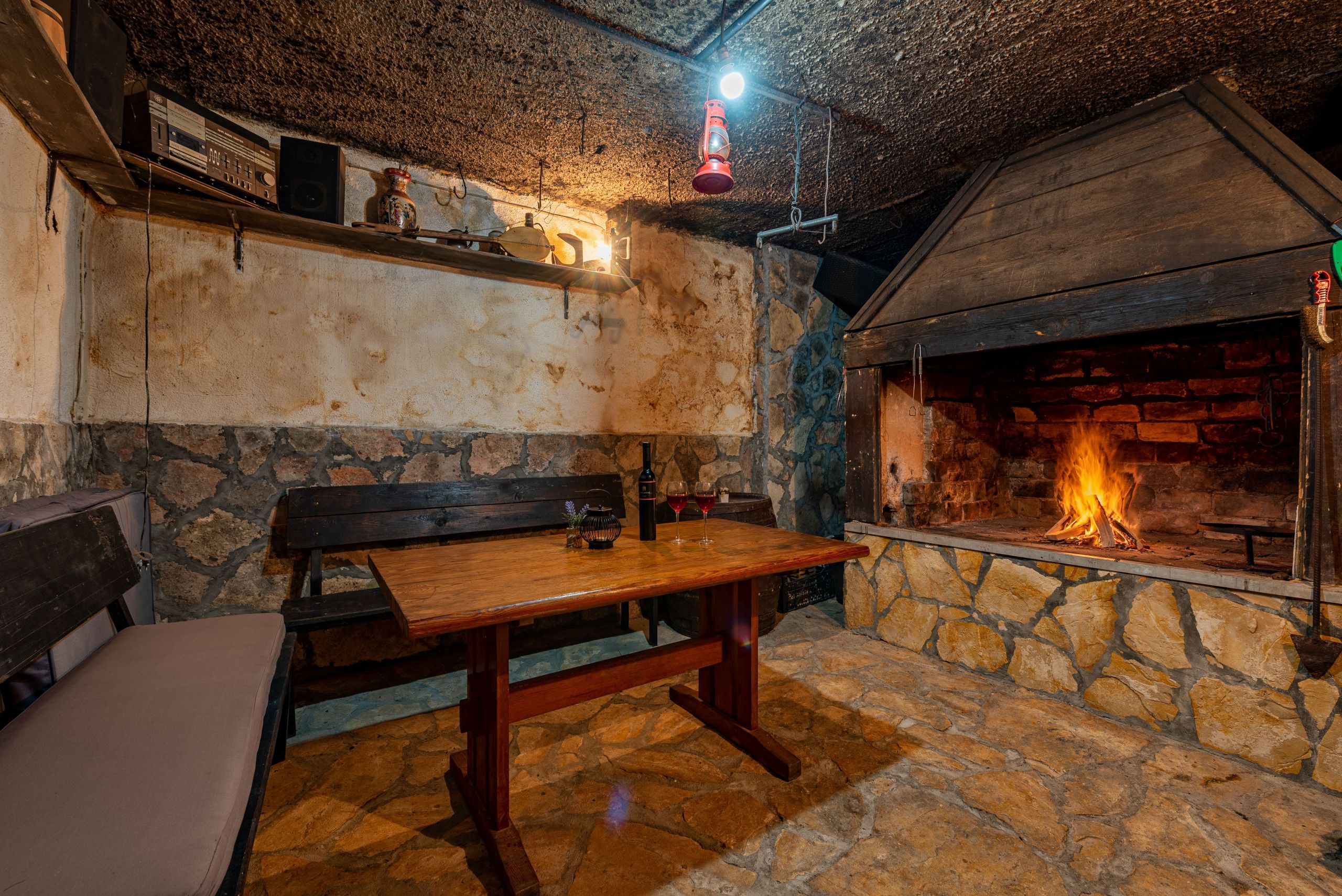 Un intérieur rustique avec un mur en pierre, une table en bois, un banc et une cheminée où l’on peut voir le feu.