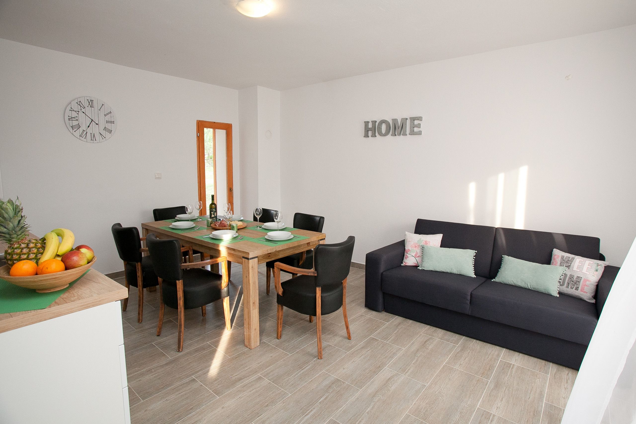 A modern living room with a dining area, featuring a wooden table, black chairs, a dark sofa with cushions, and a wall clock.