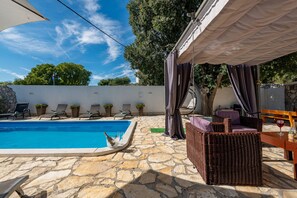 A pool area with stone flooring, lounge chairs, a wicker sofa, and a table with a glass of wine.