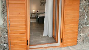 Une porte en bois donne sur une pièce dont un mur est en pierre et qui contient un lit avec une table de chevet.