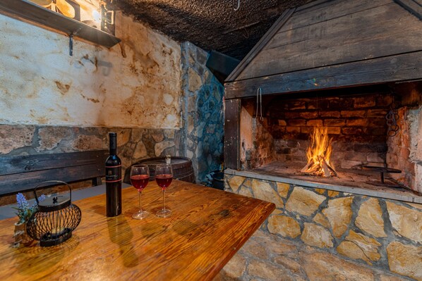 A rustic interior with a stone wall, a wooden table with a bottle and glasses, and a fireplace with a burning fire.