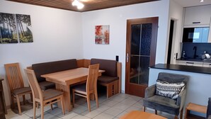 Een zithoek met een houten tafel en stoelen, een bank, en een keuken met een koelkast en spoelbak.