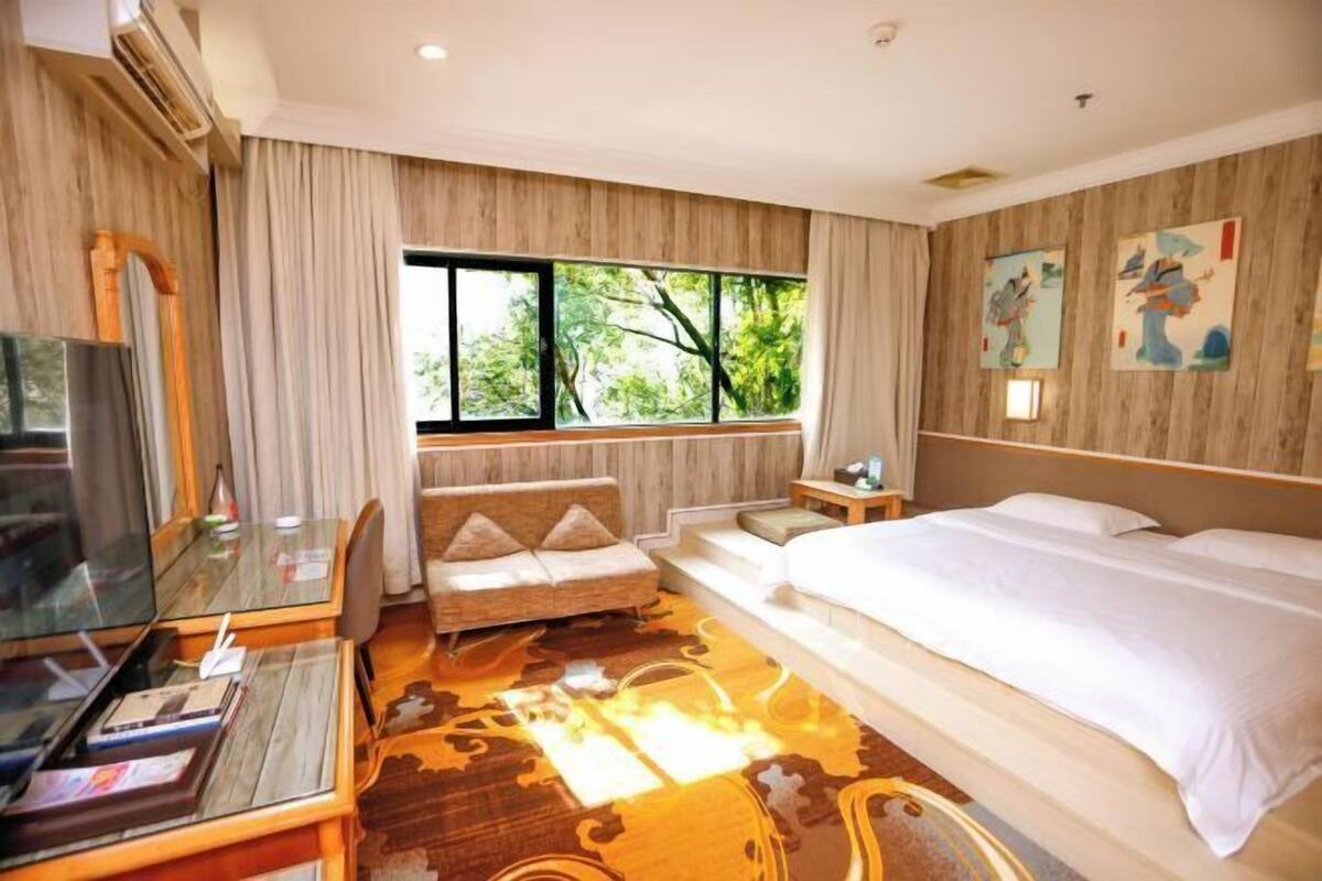 Hotelkamer met een bed, een bureau met een computer, een stoel, een bank, een televisie en een raam met uitzicht op bomen.