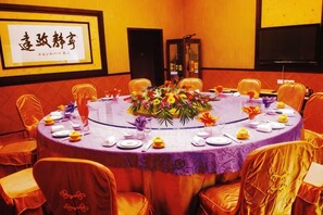Une table ronde recouverte d’une nappe violette, entourée de chaises dont les housses sont orange, dans une pièce où un panneau encadré est accroché au mur.