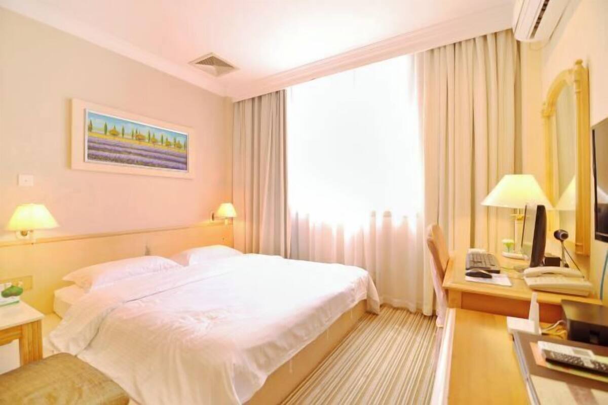 Een hotelkamer met een bed, twee bedlampen, een bureau met een computer, een stoel, een televisie en een raam met gordijnen.