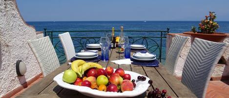 A table set with a fruit platter, wine glasses, and a bottle, overlooking the sea.
