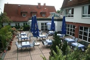 En udendørs terrasse med borde og stole, overdækket med blå parasoller, omgivet af krukker med planter.