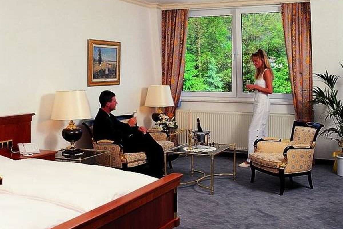 Une chambre d’hôtel comprenant un lit, deux fauteuils, une table basse et une grande fenêtre avec des rideaux.