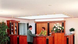 A reception desk with a man and a woman standing at it, a potted plant, and a large window in the background. - Hotel Restaurant Fasanerie (Marburg)
