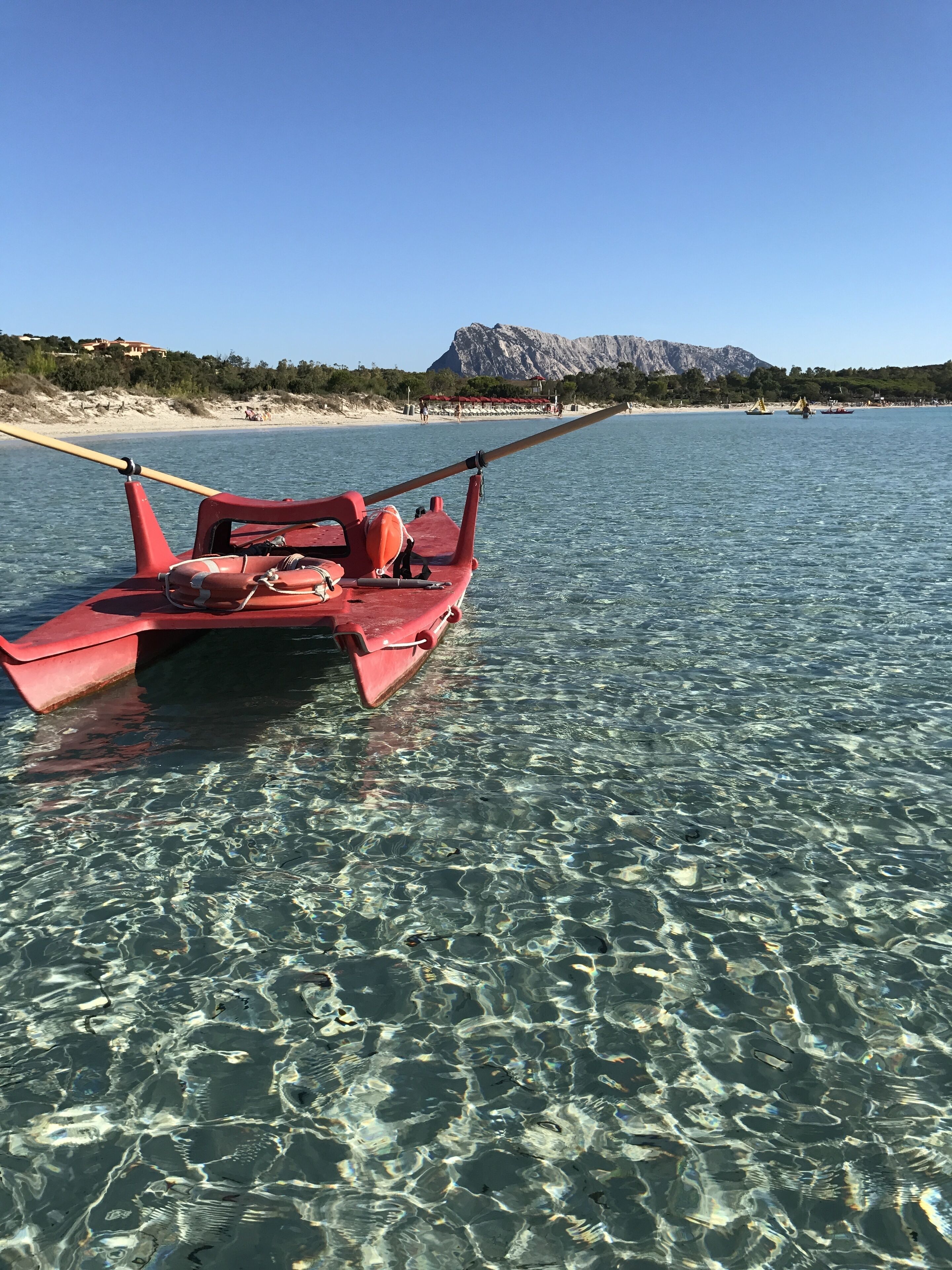 A red boat anchored near a clear beach with a mountain in the background.