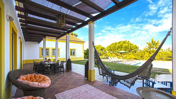 Eine überdachte Terrasse mit Gartenmöbeln, einer Hängematte und Blick auf ein Haus im Hintergrund.