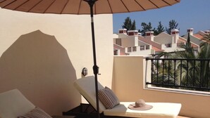 Ein sonniger Sitzbereich im Freien mit zwei Loungestühlen, einem großen Sonnenschirm und Blick auf angrenzende Gebäude.