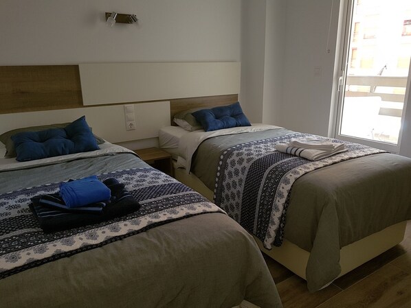 Habitación de hotel con dos camas, cada una con ropa de cama estampada y almohadas azules. Hay una ventana con persianas, una mesita de noche y una lámpara de pared.