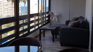 A balcony with a view of the sea and palm trees, a sofa, a table, and a lamp.