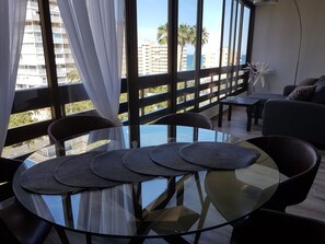 Una sala de estar moderna con una mesa de vidrio, sillas negras y vistas a palmeras y edificios a través de amplios ventanales.