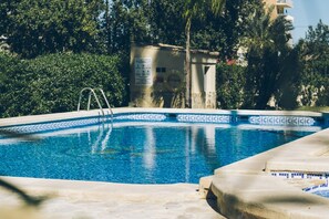 Una piscina con un borde de mosaicos azules, rodeada de árboles y un edificio al fondo.
