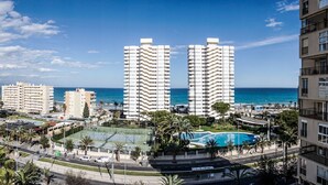 A high-rise building with a pool and palm trees in the foreground, and a beach in the background.