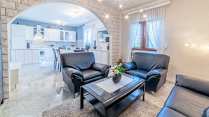 Una sala de estar moderna con sofás de cuero negro, una mesa de centro de vidrio y una cocina visible a través de un arco.