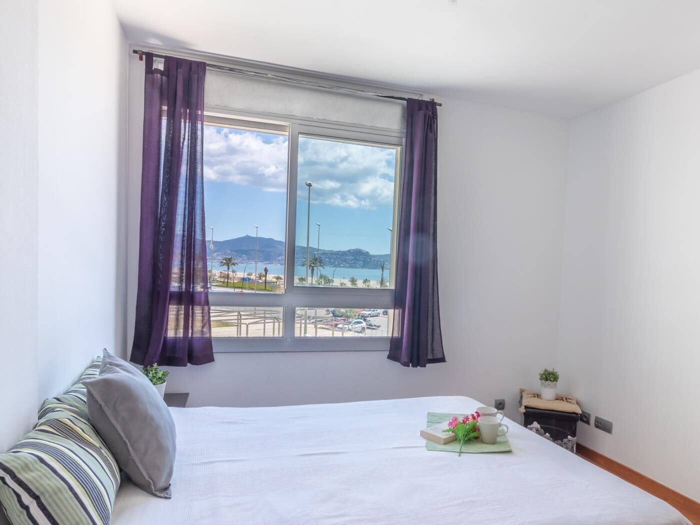 Ein Schlafzimmer mit einem Bett, einem Fenster mit Vorhängen, Blick auf Strand und Berge sowie einem kleinen Tisch mit Pflanze und Tasse.