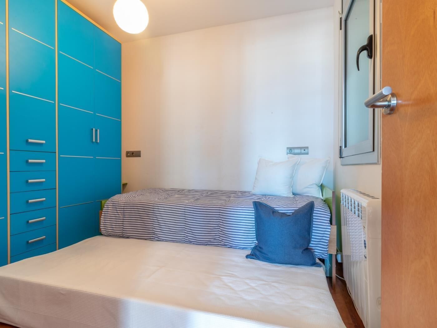 A bedroom with a bed, a blue wardrobe, a radiator, and a door with a mirror.
