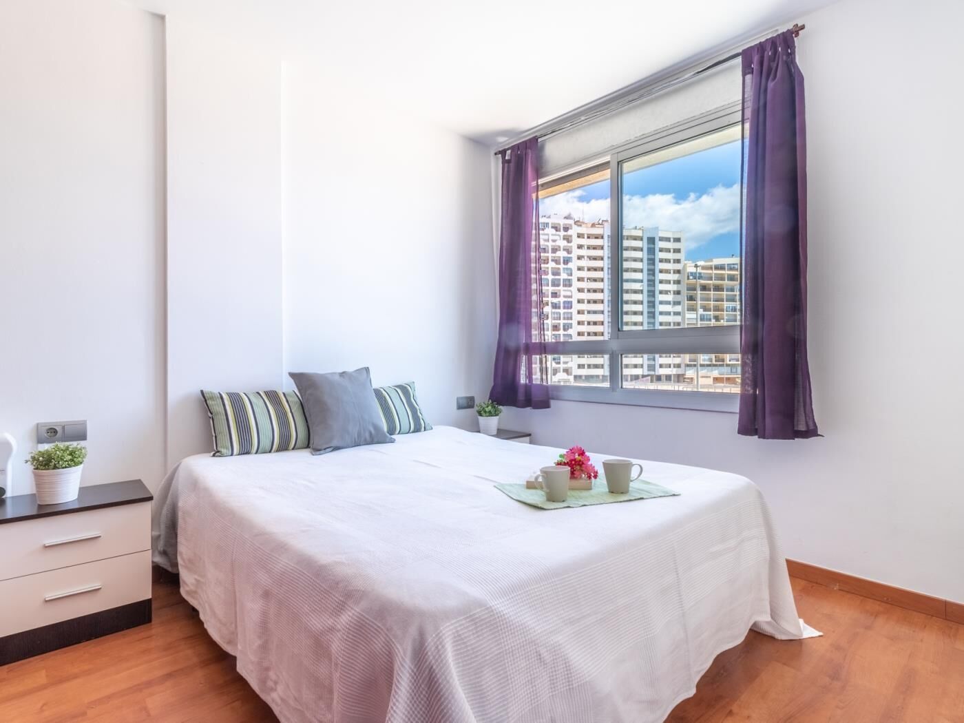 Une chambre à coucher avec un lit, une table de chevet, une fenêtre avec des rideaux et une vue sur des immeubles.