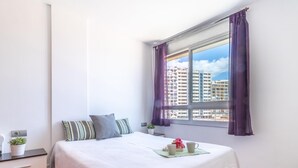 Un dormitorio con cama, mesita de noche, ventana con cortinas y vistas a edificios.