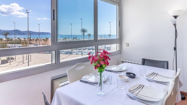 Un comedor con una mesa lista para cuatro, ventanas amplias con vistas a la playa y al mar, y un jarrón con flores rosas.
