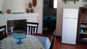 Un ambiente compatto con angolo pranzo, caratterizzato da una tovaglia a quadri e un runner da tavola in pizzo blu. Si intravede un frigorifero bianco alla destra, e un letto sullo sfondo.