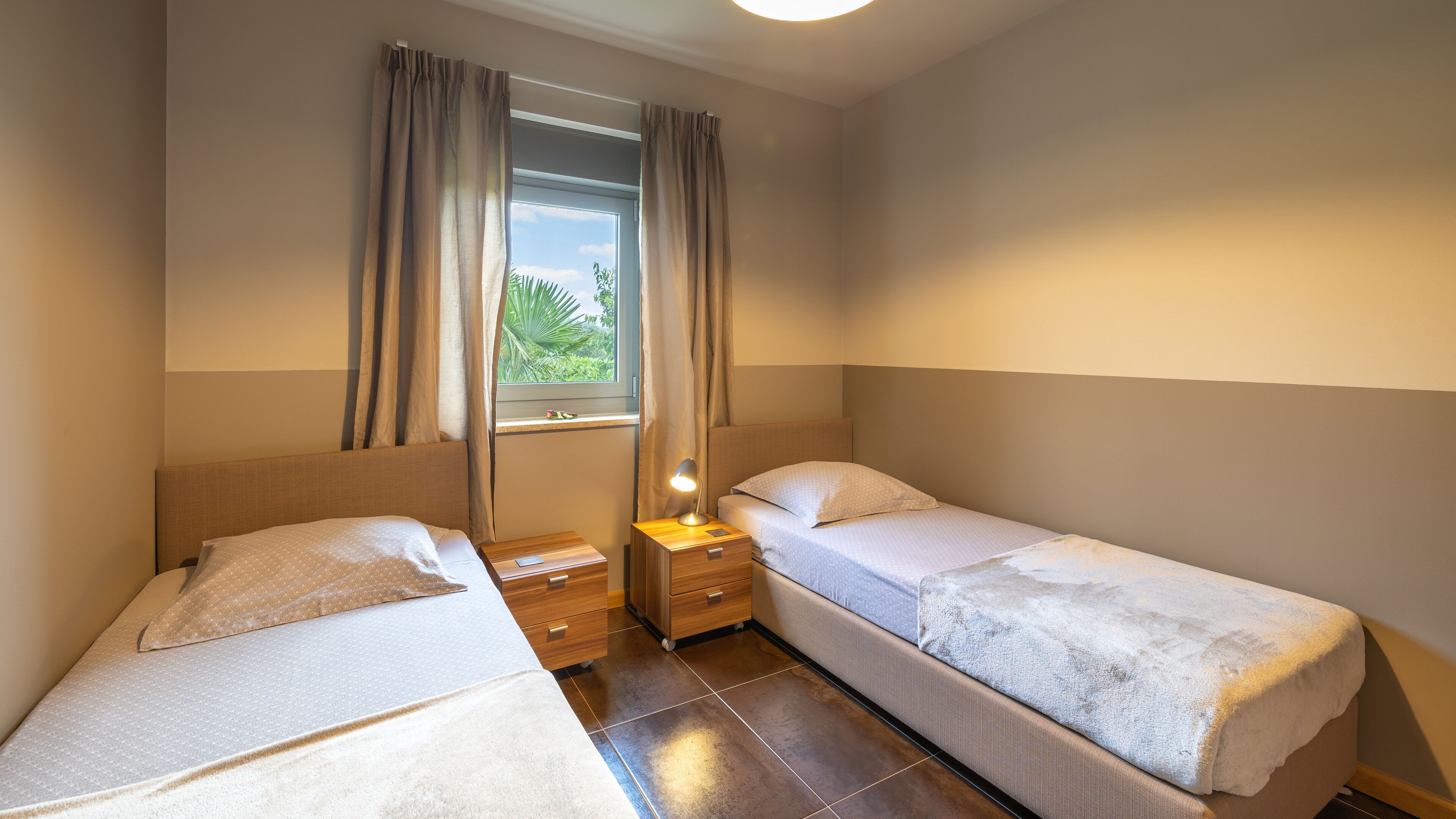 A twin room with two beds, a window with curtains, a bedside table with a lamp, and a view of greenery outside.
