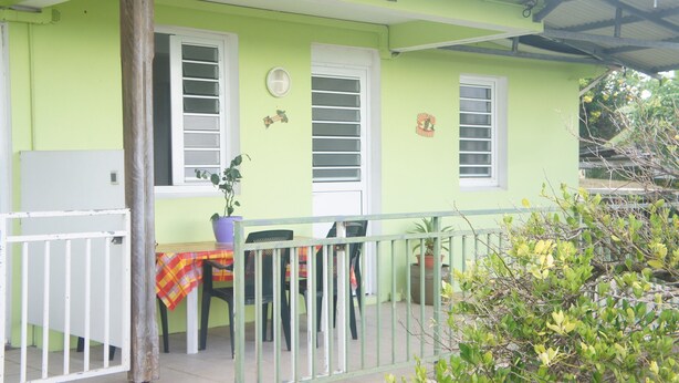 Uma casa verde vibrante com varanda, uma mesa de jantar com cadeiras e um vaso de planta.