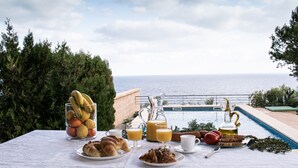 Ein Frühstückstisch mit Croissants, Obst und Getränken mit Blick auf einen Pool und das Meer.