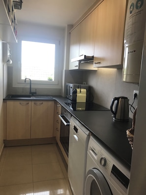 Cocina compacta con armarios de madera, microondas integrado, tostadora, tetera y lavadora.