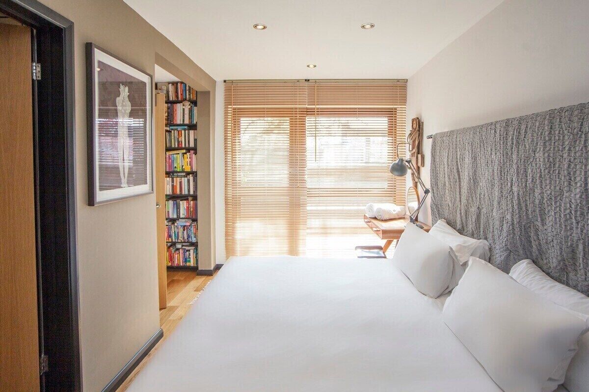 A bedroom with a large bed, a bookshelf, a framed picture, and a window with blinds.