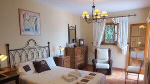 Un dormitorio bien iluminado con cama, mesitas de noche, cómoda, silla y ventana con cortinas.