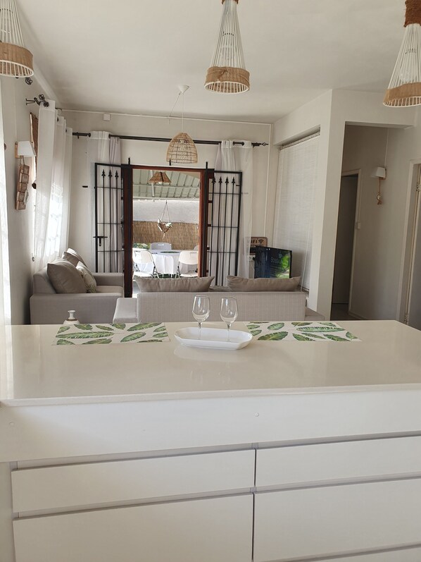 Un salon moderne avec un canapé, une table à manger agrémentée d’un plateau et de verres, et une vue sur un espace extérieur aménagé avec des tables et des chaises.